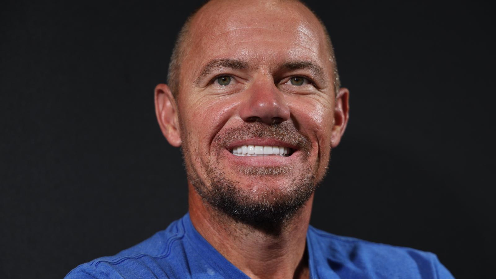 A bald man with a light beard and blue shirt is smiling against a dark background.