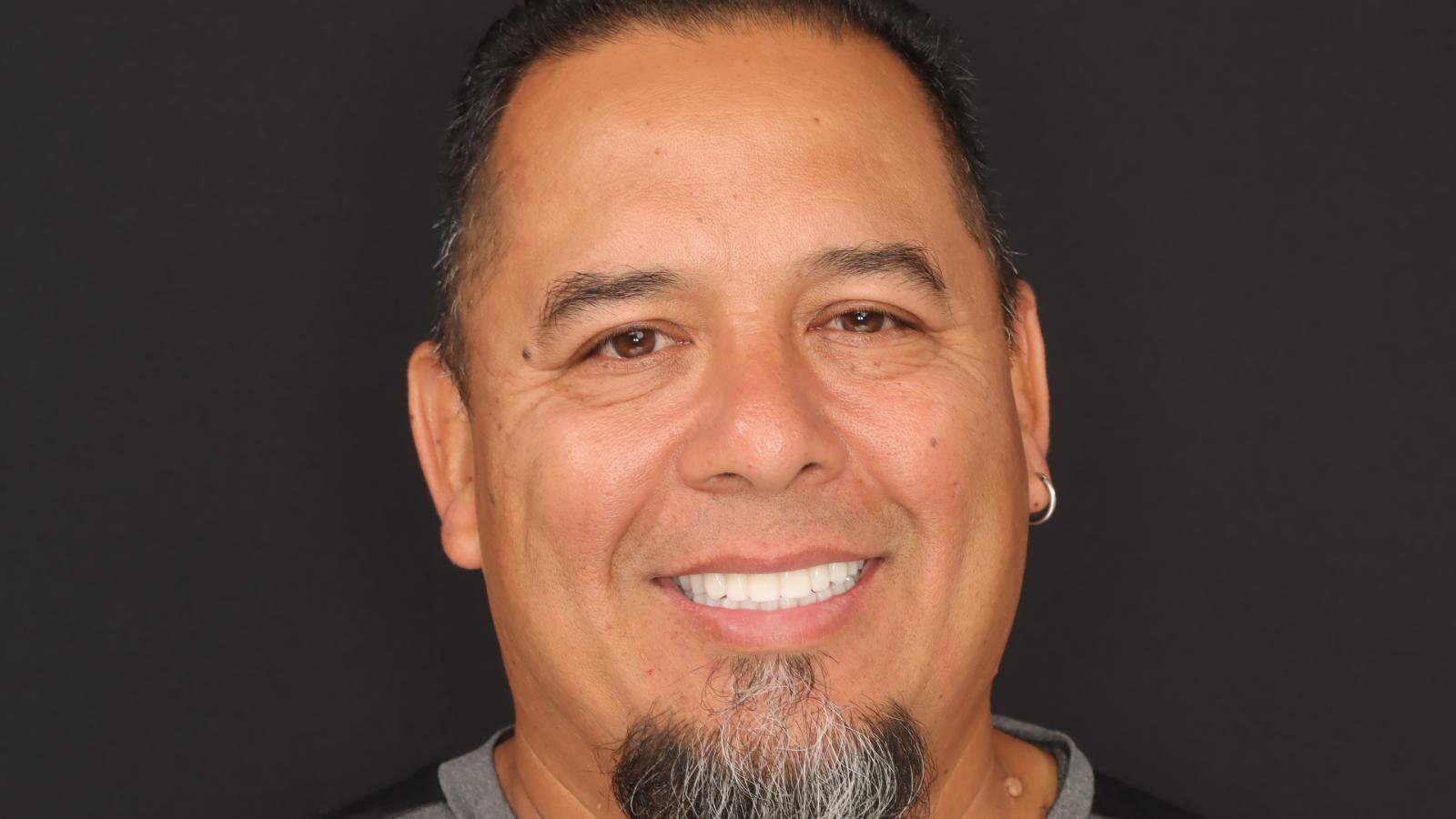 A man with short dark hair, a smile, and a goatee is photographed against a plain black background. He is wearing a black shirt.