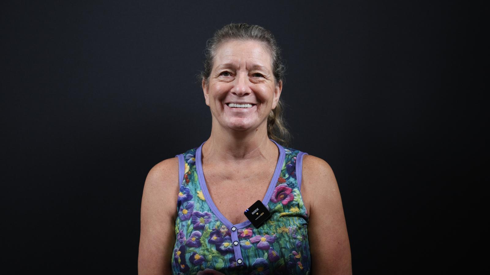 A woman with tied-back hair smiles at the camera, wearing a floral tank top and a small black microphone clipped to her shirt against a plain black background.