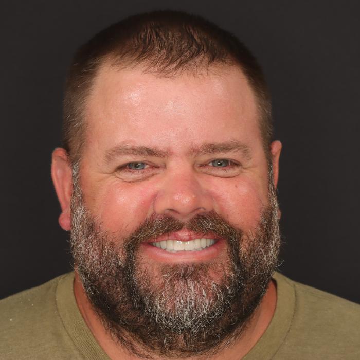 A bearded man with short hair smiles at the camera against a black background.