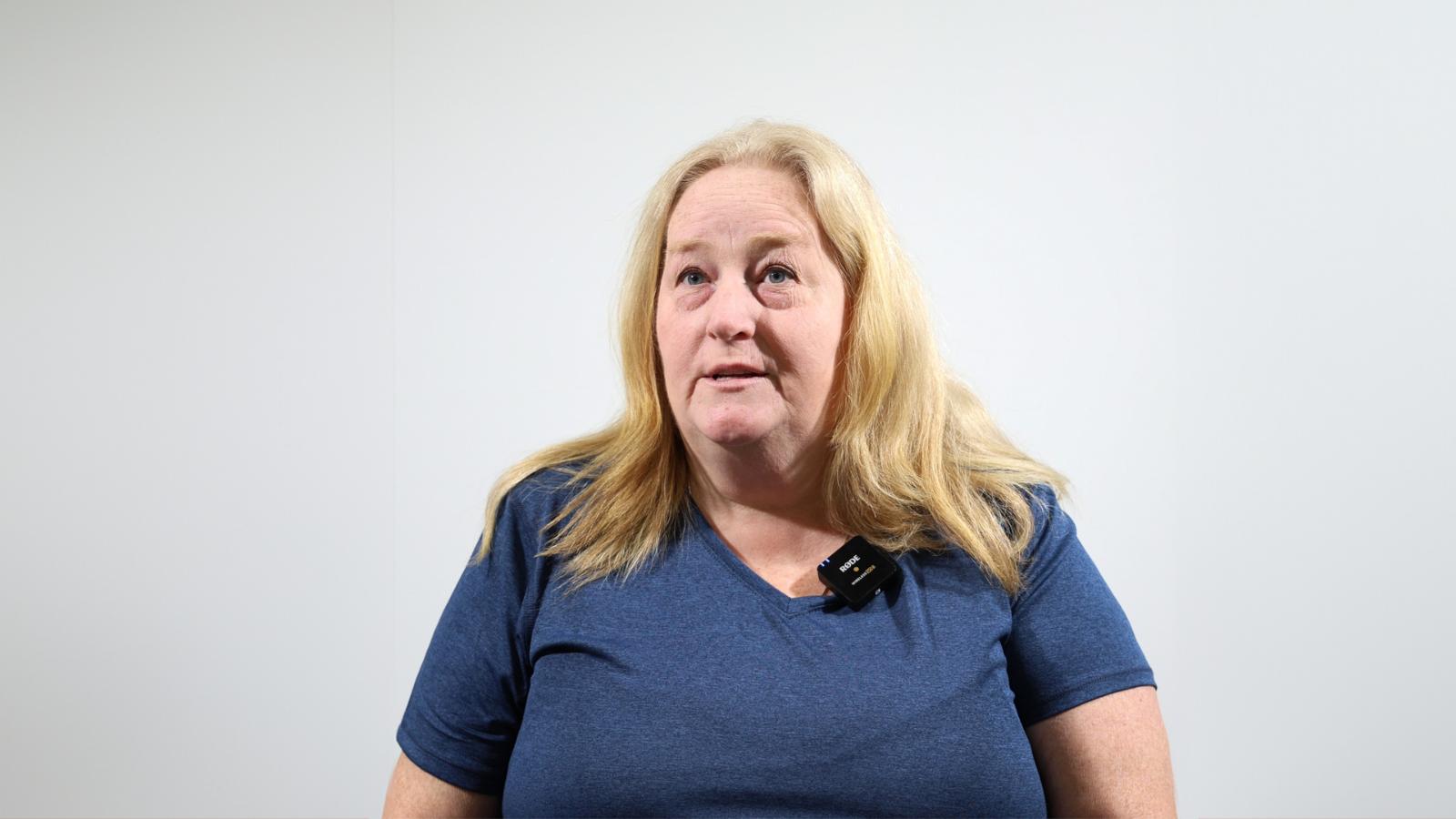 A woman with long blonde hair wearing a blue shirt and a microphone clipped on it stands against a plain white background.