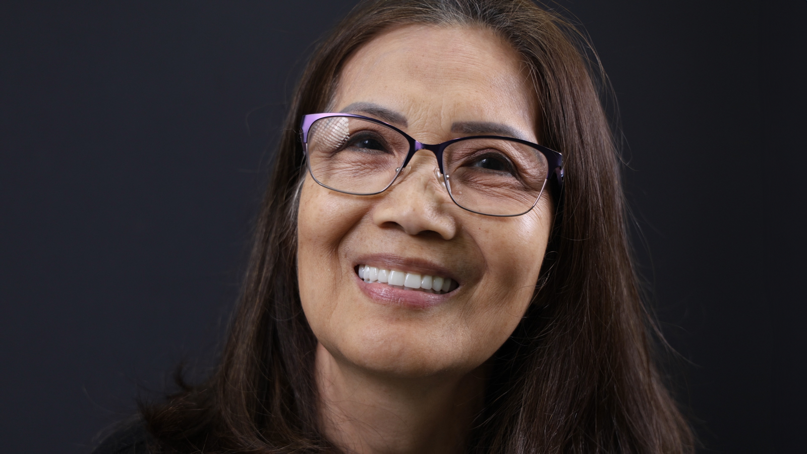 A woman with long brown hair and glasses smiles while looking upward against a dark background.