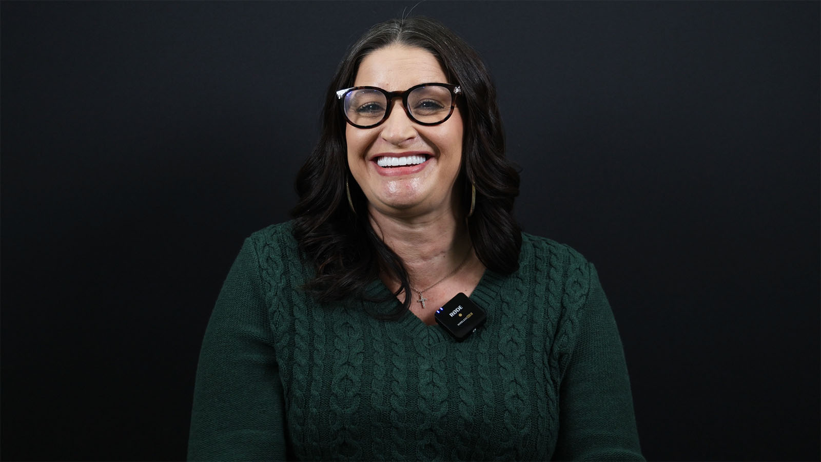 A woman with long dark hair and glasses smiles in front of a black background, wearing a green sweater and a necklace.