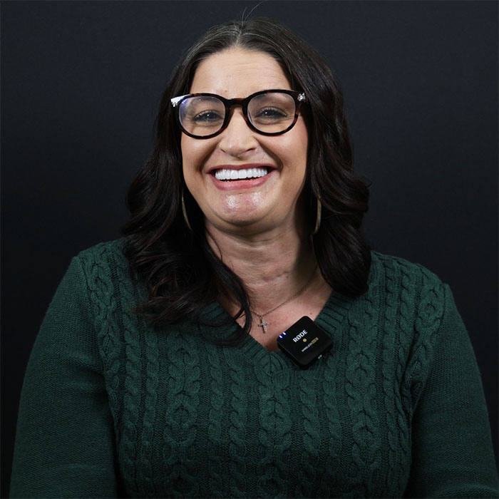 A woman with glasses and long dark hair is smiling, wearing a green sweater and a microphone clipped to her shirt. The background is black.