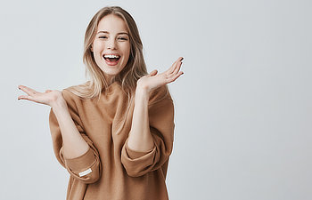 A person with long hair smiles and raises both hands, appearing cheerful, against a plain background. They wear a light brown sweater.