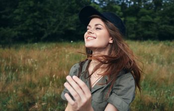 A person with long hair and a black cap smiles outdoors in a grassy area with trees in the background.