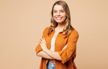 Young woman with long hair and a confident smile, wearing an orange shirt and light jeans, stands with arms crossed against a plain beige background.