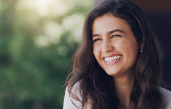 Woman with long dark hair smiling outdoors with blurred greenery in the background.