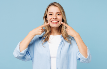 A smiling young woman in a light blue shirt points to her teeth with both index fingers against a blue background.