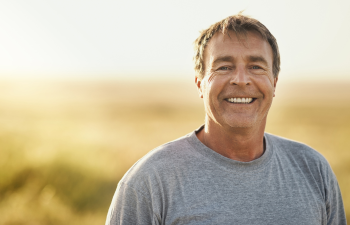 Middle-aged man smiling in a gray t-shirt, standing outdoors with a blurred natural background.