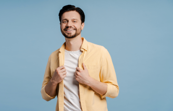 A man in a yellow shirt stands against a blue background, smiling and holding both sides of his open shirt.