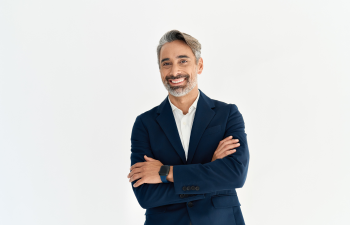 Smiling man with gray hair and a beard, wearing a dark suit and white shirt, stands with arms crossed against a plain white background.