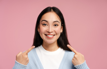 Smiling person with long dark hair points at themselves, wearing a blue cardigan and white shirt, set against a pink background.