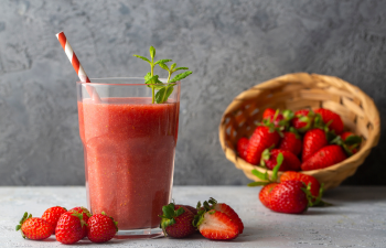 A glass of strawberry smoothie with a red and white straw and mint garnish is placed next to strawberries and an overturned basket on a gray surface.