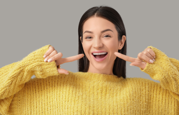 Person smiling and pointing at their teeth, wearing a yellow sweater, against a gray background.