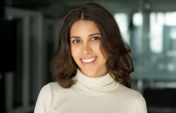 A person with long brown hair smiles while wearing a white turtleneck sweater. The background is softly blurred.