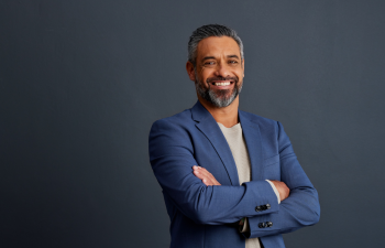 Man with gray hair and beard smiling, arms crossed, wearing a blue blazer over a light shirt against a dark background.